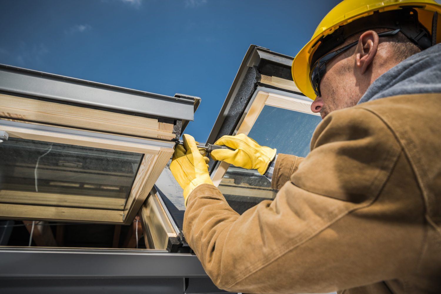 Caucasian Worker Wearing Safety Helmet and Protective Gloves Carrying Out Repair Works on Roof Skylight Windows Using His Screwdriver.